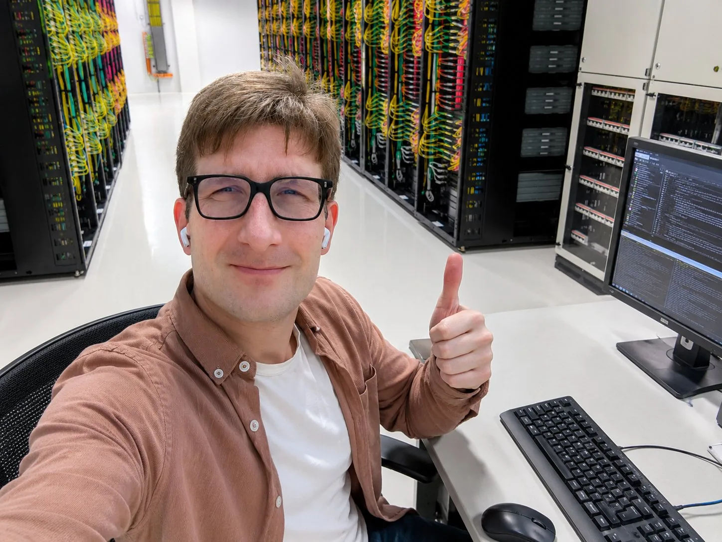 A smiling man in glasses takes a thumbs-up selfie from a desk inside a datacenter aisle lined with GPU racks full of colorful cabling.